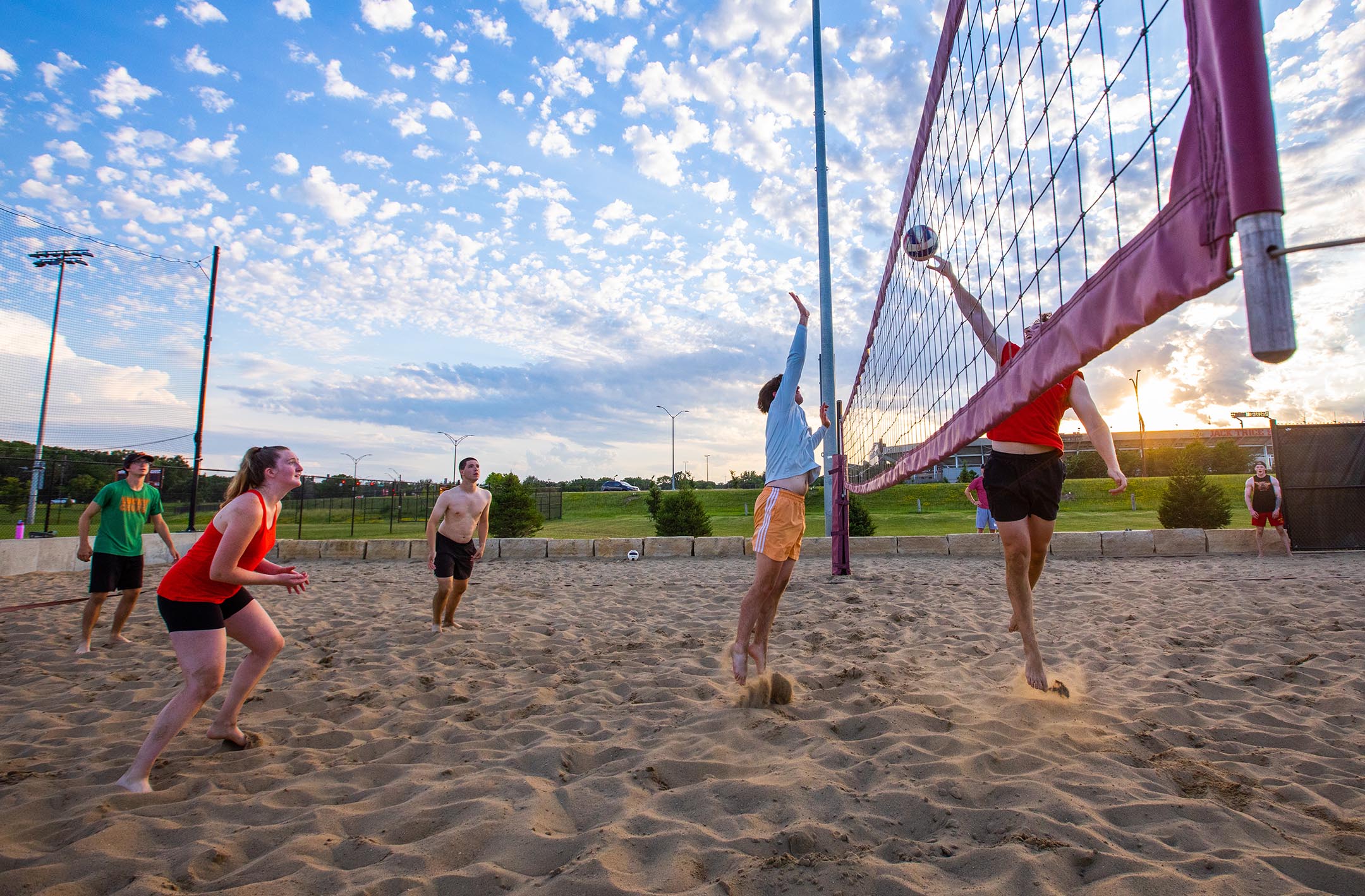 Students playing sand volleyball at sunset with Jack Trice Stadium in the background.