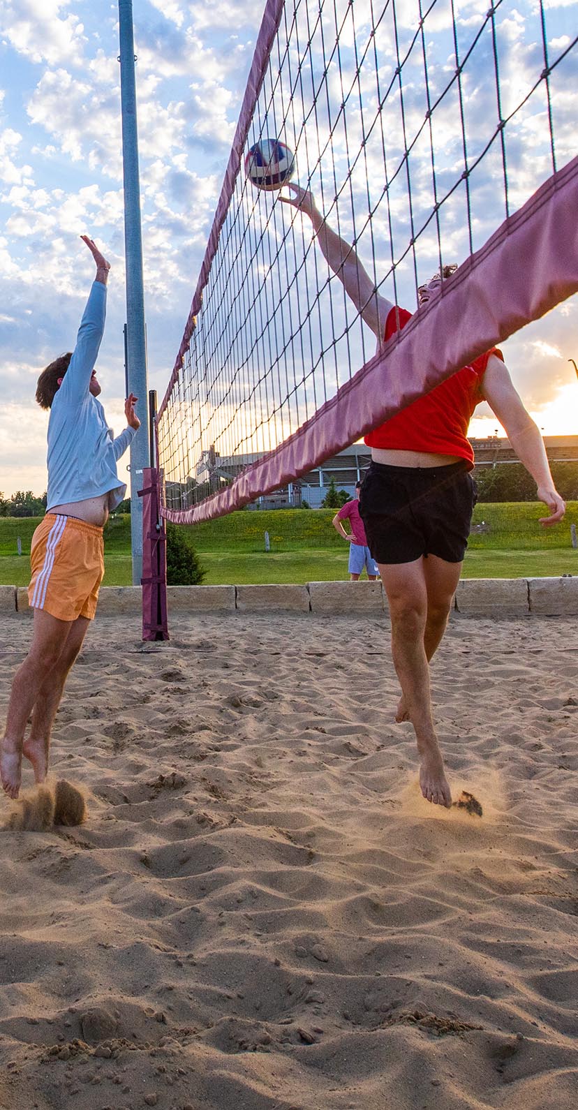 Students playing sand volleyball at sunset with Jack Trice Stadium in the background.