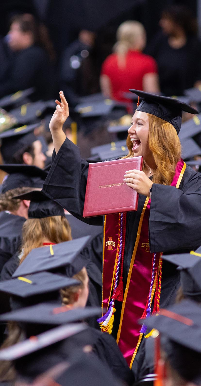 A graduate holding up a diploma and waving into the crowd at commencement.