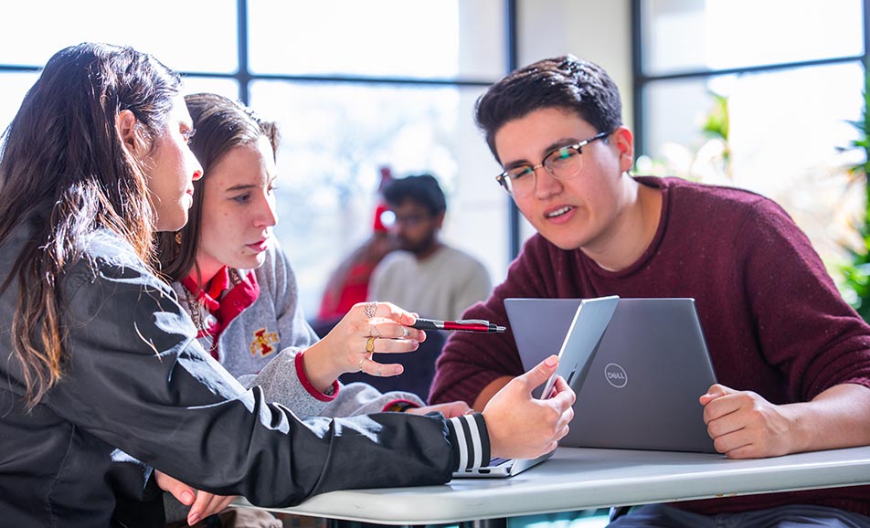 Three students talking while seated at a study table in the library.